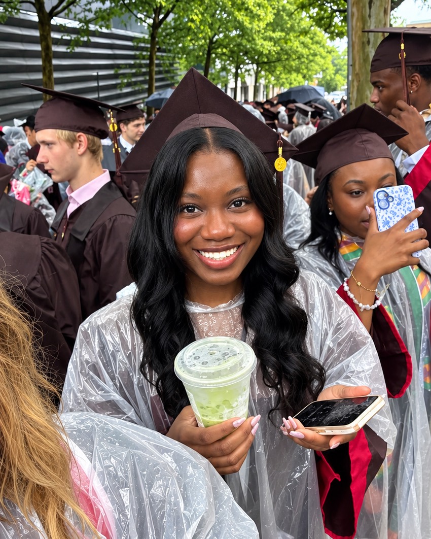 Tomi smiling at her Brown University graduation in her cap and gown, under a rain poncho